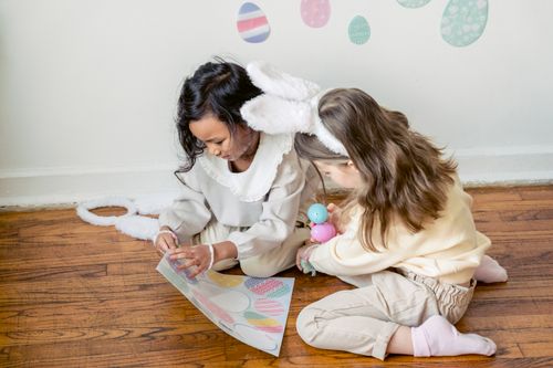 Free From above of concentrated little multiethnic friends in stylish clothes and bunny ears ripping stickers from paper sheet while decorating wall before Easter celebration Stock Photo