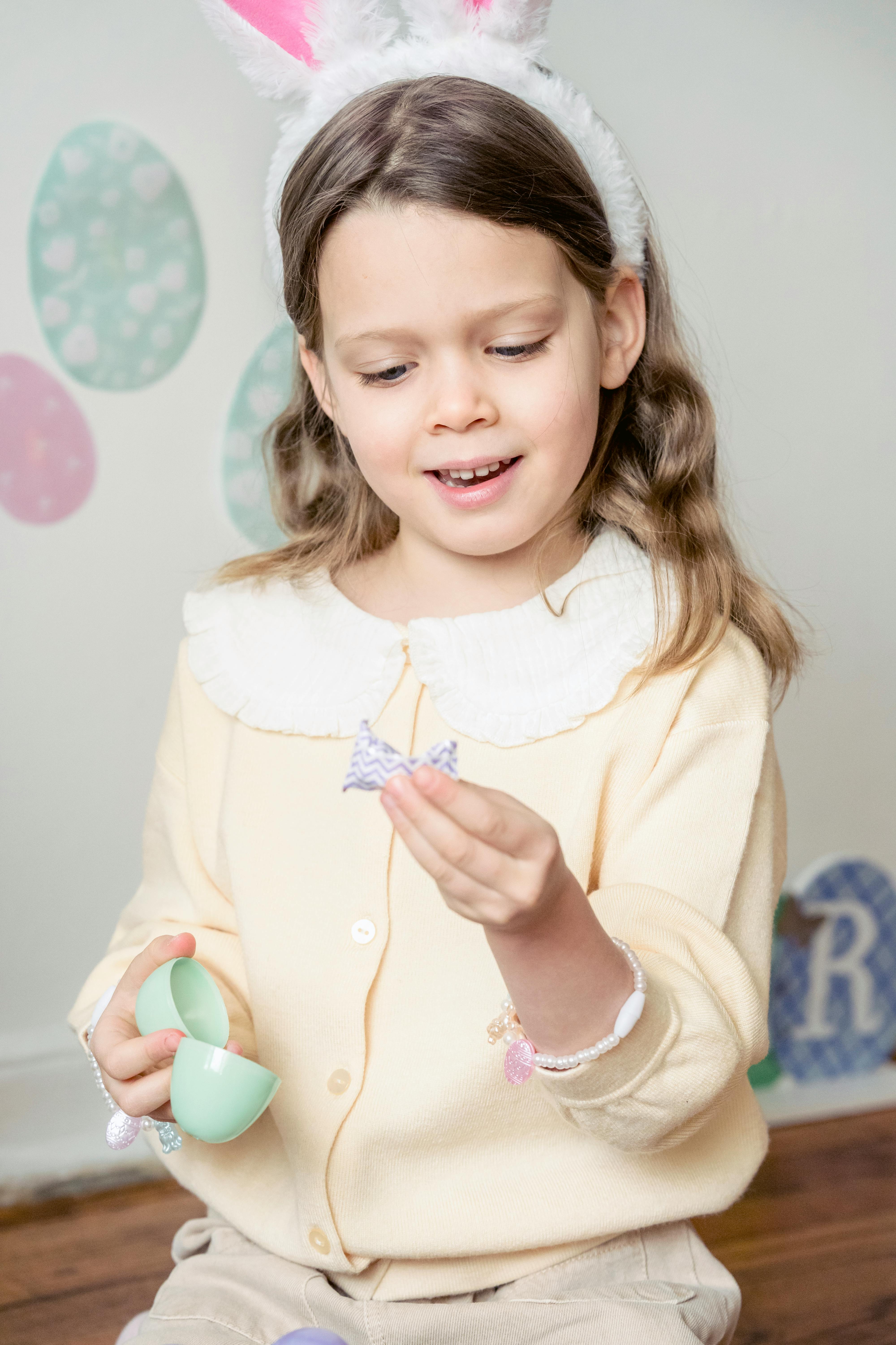 Excited little girl with long wavy hair in casual clothes and bunny ear headband looking at wrapped candy found in egg toy during Easter celebration