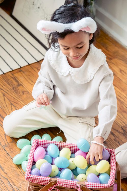 Free From above of adorable little Hispanic kid with bunny ears sitting on wooden floor and playing with colorful artificial Easter eggs placed in wicker basket Stock Photo