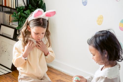 Free Cute calm little girl with bunny ears on head sitting on floor at home and playing with ethnic friend during Easter holidays Stock Photo