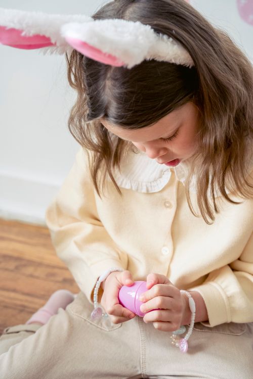 Free From above of adorable serious little girl with long wavy hair in stylish clothes and bunny ears headband sitting on wooden floor and opening pink egg toy during Easter holidays Stock Photo
