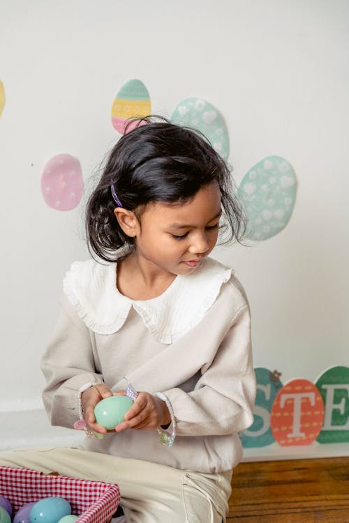 Little Hispanic girl playing with plastic eggs near decorated wall