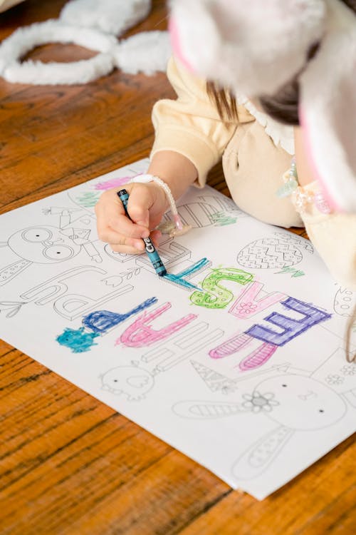 From above of crop anonymous little girl sitting on wooden floor and drawing in coloring book in daylight