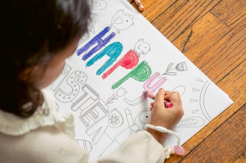 Free From above of crop anonymous kid lying on floor and drawing inscription Happy Easter on coloring book in daytime Stock Photo
