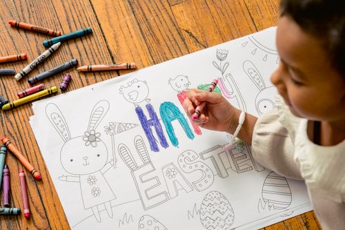 Free From above of crop ethnic girl painting in coloring book with Happy Easter inscription at home in daytime Stock Photo