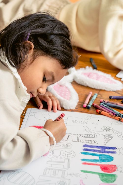 Free Side view of focused ethnic girl lying on floor and drawing in coloring book at home in daylight Stock Photo