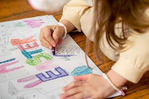 Free High angle of crop unrecognizable kid in casual clothes sitting on wooden floor and coloring paper with Easter word and eggs with wax pencil in hand in light room Stock Photo
