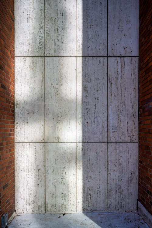 Wall with weathered white wooden panels between brown brick walls of building located on street with bright sunlight in town