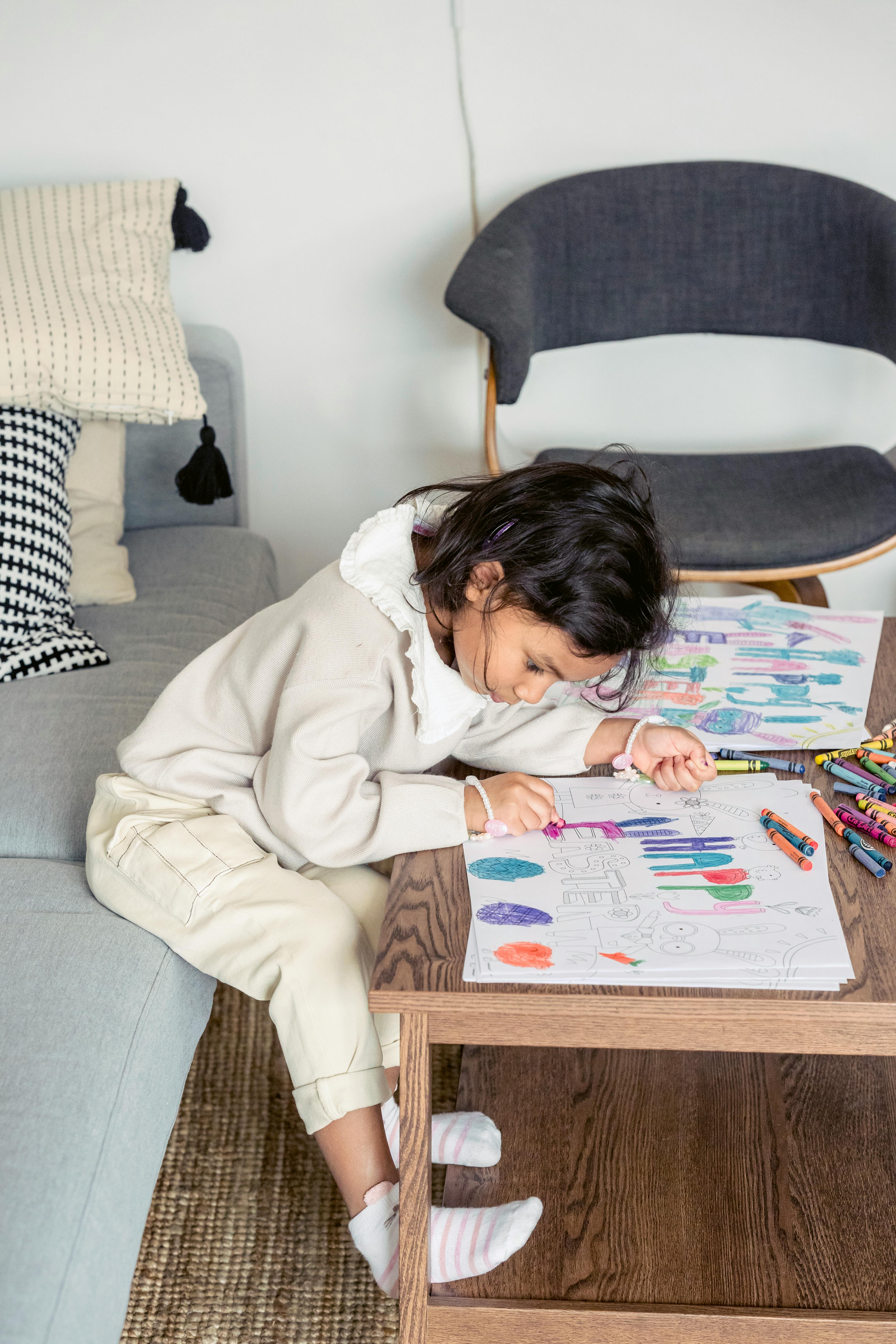 Free Side view of Hispanic girl sitting on couch at table and coloring picture with wax pencils for Easter Stock Photo