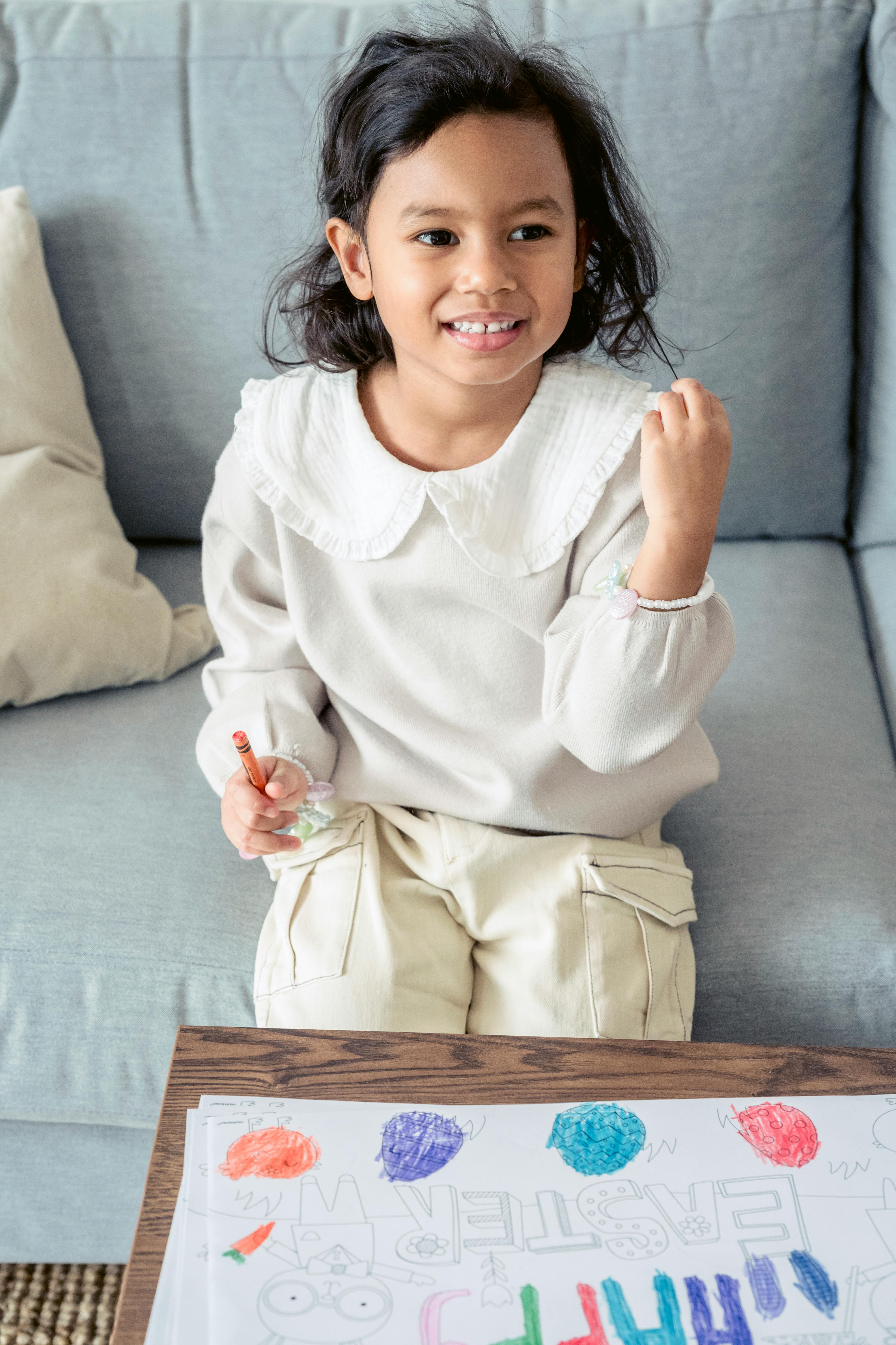 High angle of positive Hispanic girl sitting on couch and coloring Easter picture for Easter celebration