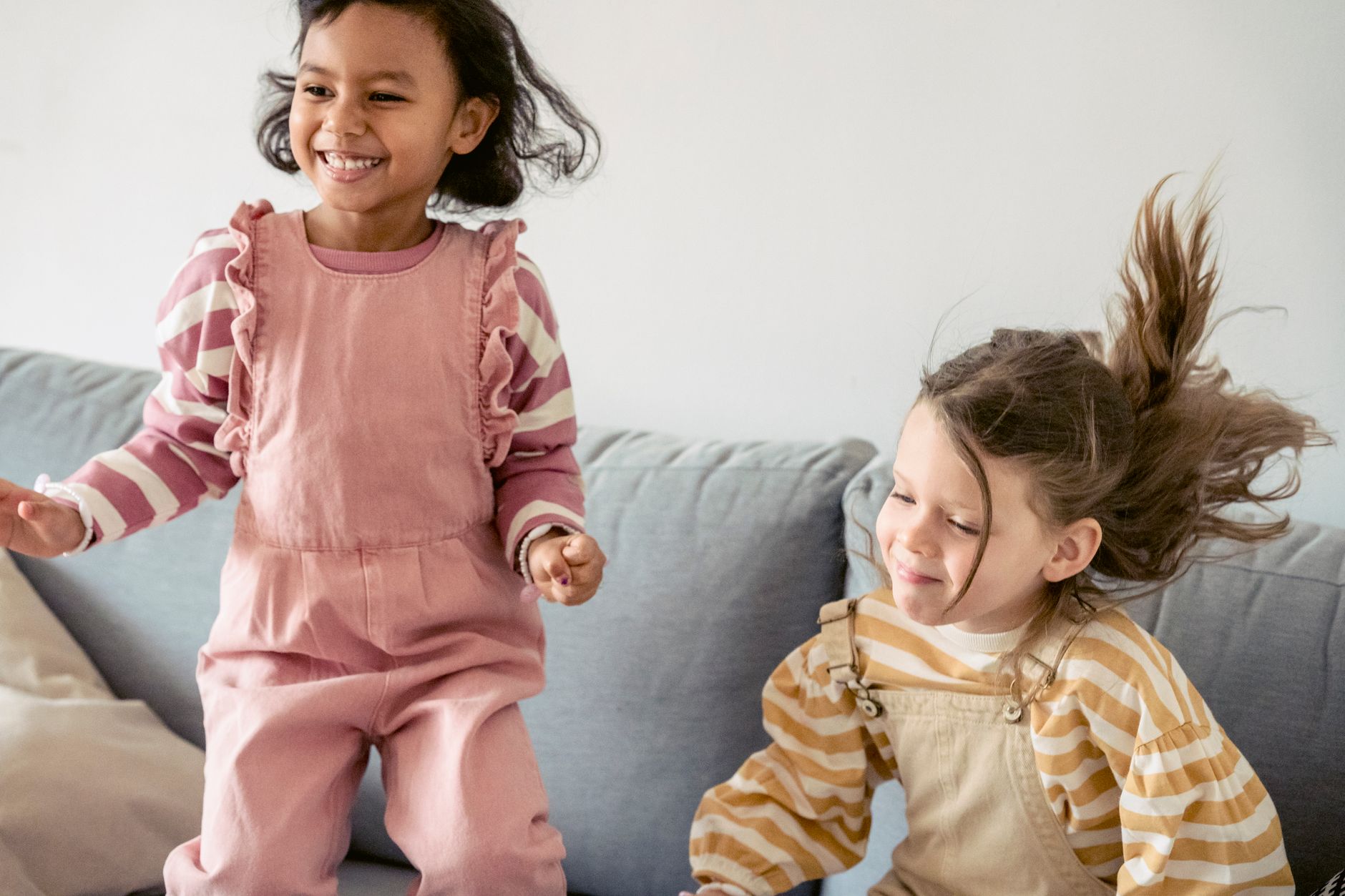 Cute diverse little girls jumping on sofa and smiling happily while spending pleasant time together