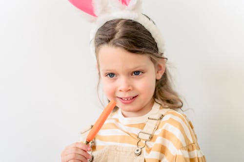 Cute little girl wearing bunny ears touching face with carrot during Easter holiday and smiling against white background
