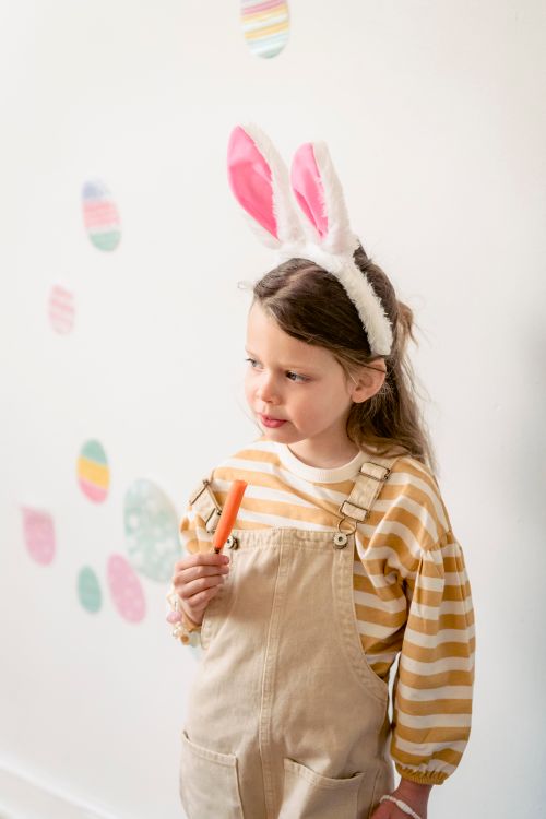 Free High angle of adorable preschool girl wearing bunny ears chewing peace of carrot while standing against white wall with Easter decor Stock Photo