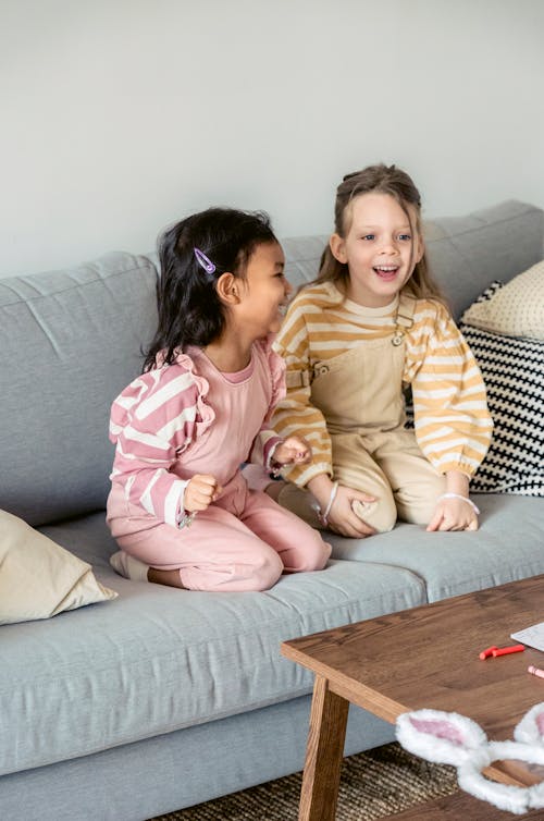 Full body of happy multiethnic girls laughing happily while sitting on couch near table with crayons and bunny ears