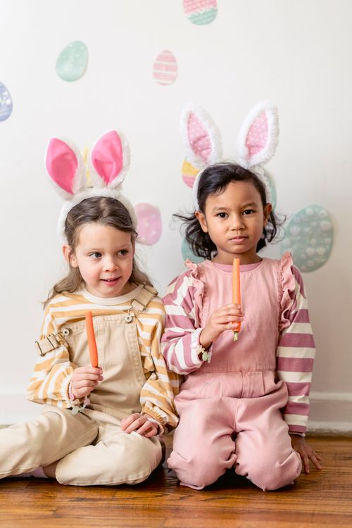 Free Cute little multiethnic friends in similar clothes and bunny ears sitting together with carrots against wall with Easter eggs stickers Stock Photo