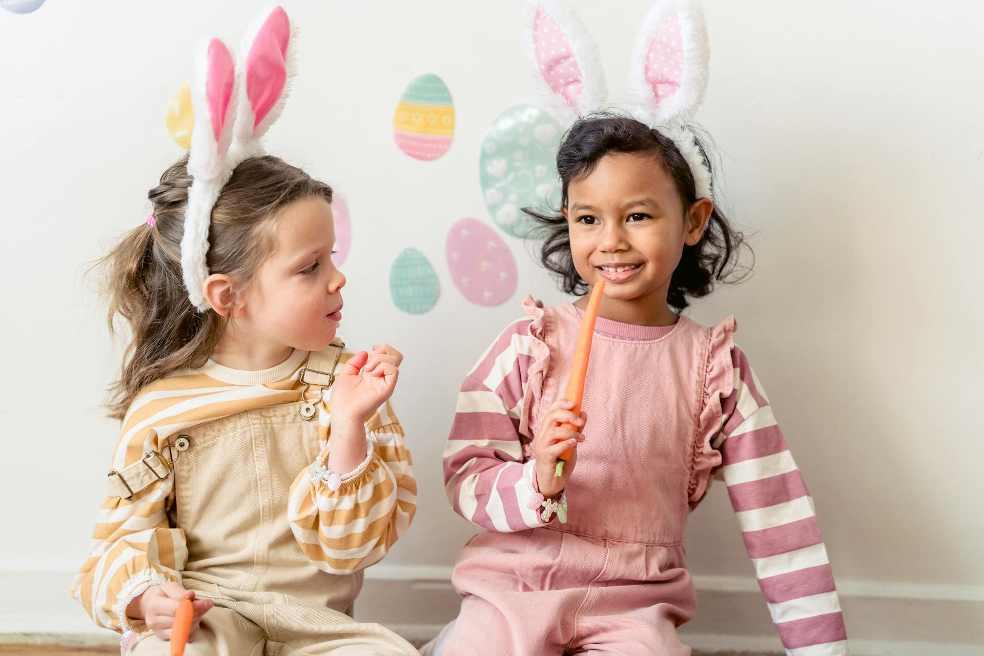 Adorable Hispanic girl with carrot sitting near friend in bunny ears against wall decorated with egg stickers