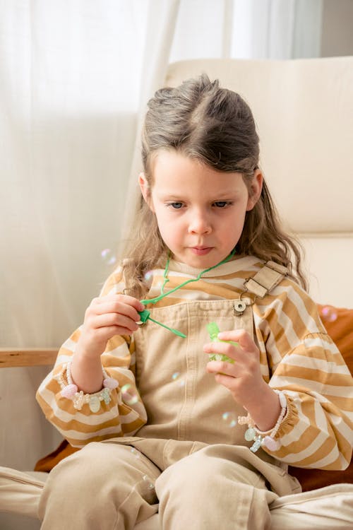 Free Charming girl in striped sweatshirt and overall sitting in armchair with soap bubbles in hands Stock Photo