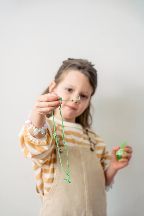 Free Cute little preschool girl in overall demonstrating soap bubble while standing against white wall Stock Photo