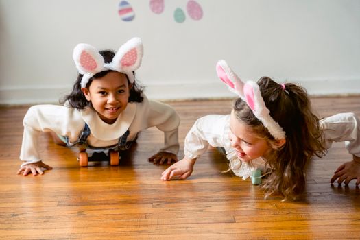 Two children wearing bunny ears playfully crawl on a wooden floor, embracing Easter joy.