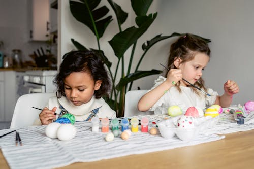 Concentrated multiracial girls painting eggs with paintbrushes while sitting at table with set of paints in kitchen during Easter holiday preparation