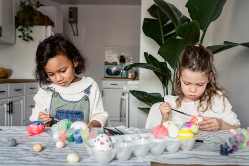 Free Concentrated diverse preschool girls with paintbrushes painting eggs at table with collection of paints in light kitchen with plant during Easter holiday Stock Photo