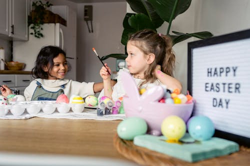 Content preschool multiracial girls with paintbrushes painting eggs at table with signboard with Happy Easter Day inscription in light kitchen