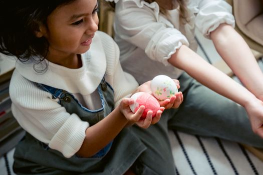 Cheerful Hispanic girl with creative colorful eggs sitting near anonymous kid in light room during Easter holiday celebration at home
