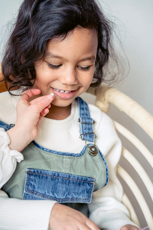 Positive preschool Hispanic girl sitting near crop anonymous friend with decorative stickers while playing together in light room at home