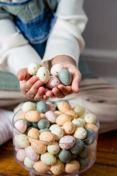 Free Crop unrecognizable kid handful with quail eggs sitting on floor near glass bowl full of small colorful eggs during Easter celebration Stock Photo