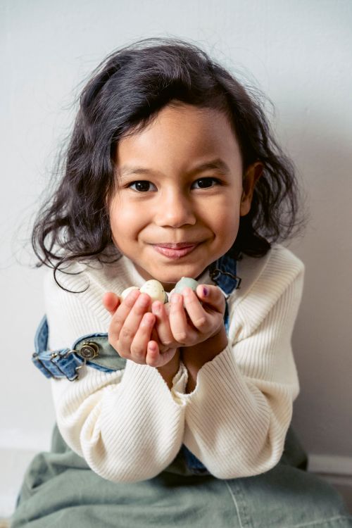 Free Cheerful preschool Hispanic girl handful of colorful quail eggs looking at camera while sitting on white background during Easter celebration Stock Photo