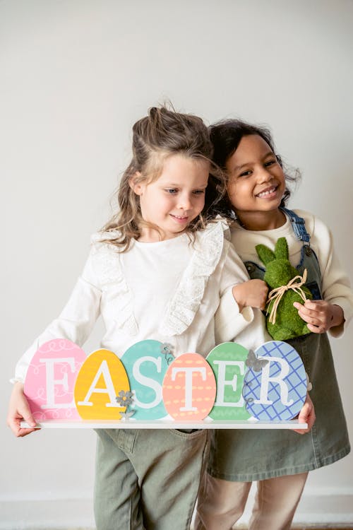 Positive multiethnic girls with bunny and egg decoration forming Easter word standing on white background during holiday celebration in room