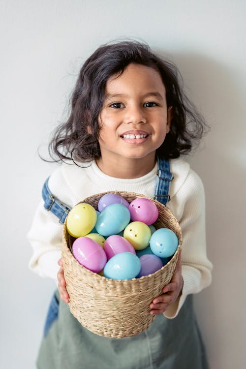 Free Adorable Hispanic girl with wicker basket full of colorful eggs looking at camera on white background during Easter holiday celebration Stock Photo