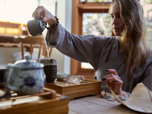 Free A woman elegantly pours tea from a pot into a cup on a wooden tray, creating a serene setting. Stock Photo