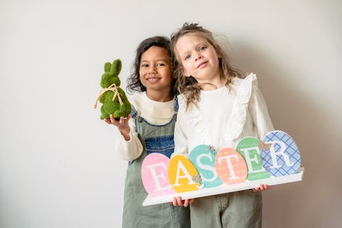 Positive multiethnic girls with decorative bunny and wooden egg decoration forming Easter word looking at camera on white background during holiday celebration