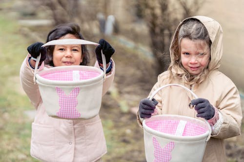 Cheerful multiracial girls with baskets in hands looking at camera while playing in backyard on blurred background during Easter holiday