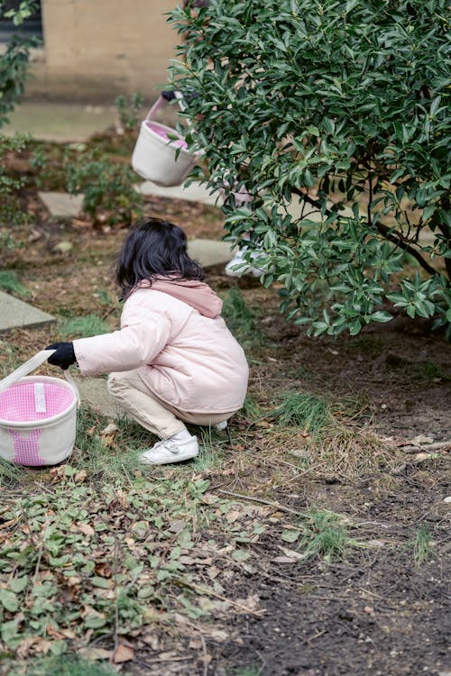 Free Back view anonymous girl in outerwear carrying soft bucket and hunkering down in spring garden during Easter hunt Stock Photo