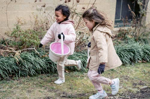 Free Full body glad diverse girls wearing warm clothes carrying soft buckets and searching for Easter eggs together in spring garden Stock Photo