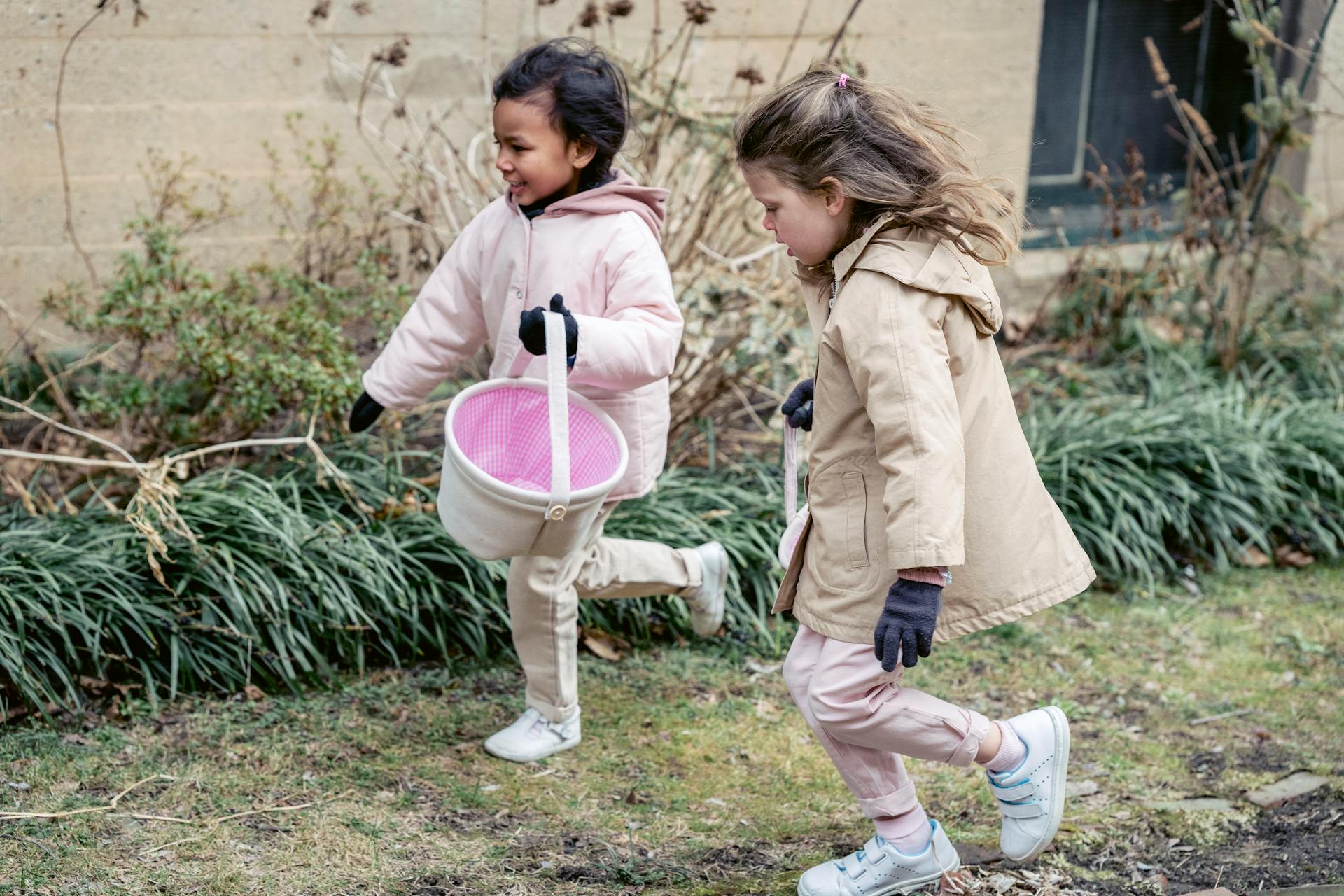 Full body glad diverse girls wearing warm clothes carrying soft buckets and searching for Easter eggs together in spring garden