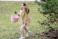 Full length cheerful girl in warm clothes carrying fabric basket and demonstrating ripe green colored egg or Easter egg in spring park