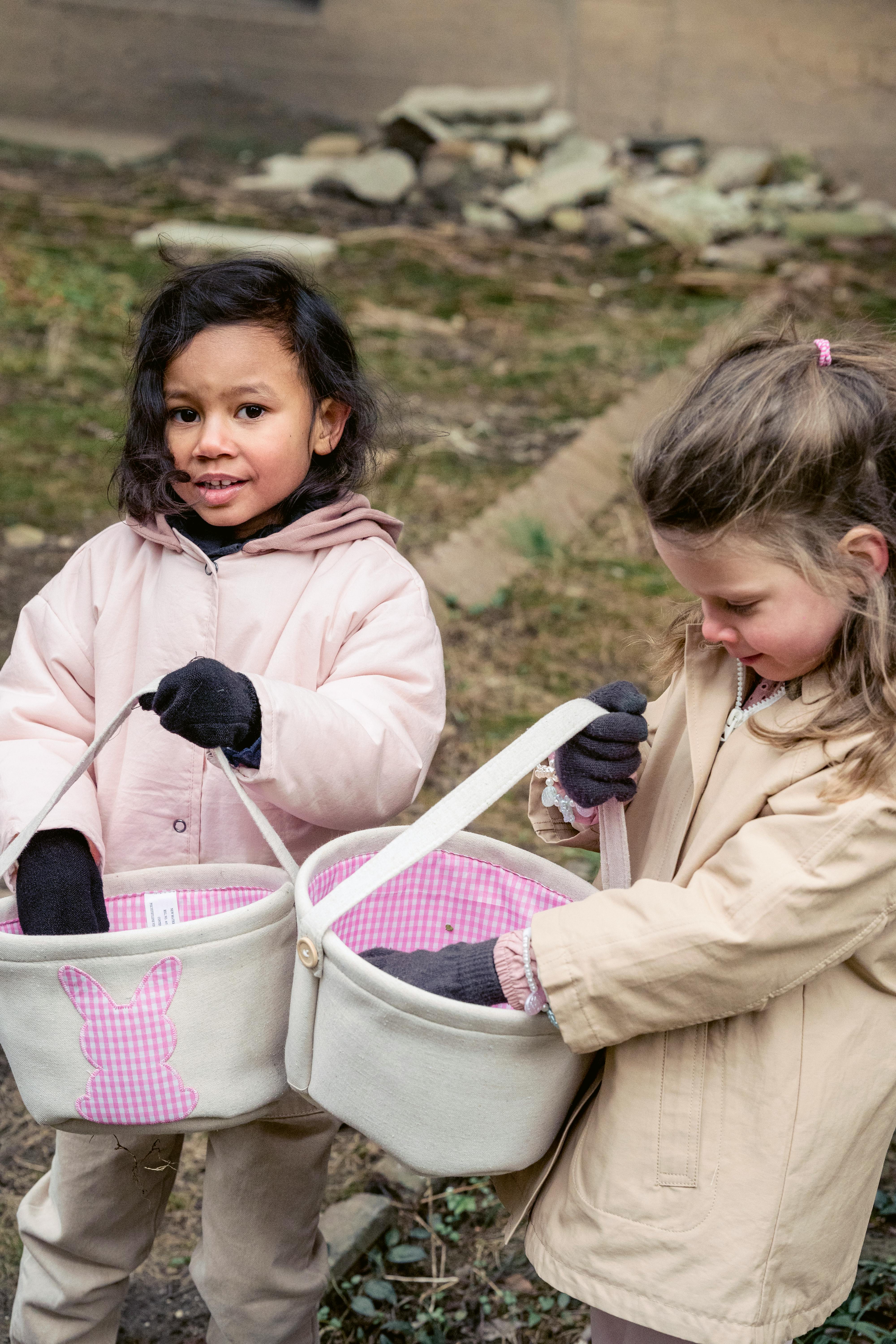 Positive multiracial girls wearing warm clothes collecting harvest in fabric buckets while standing in spring garden