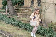 Full length girl in warm clothes carrying fabric bucket and searching for hidden Easter eggs while walking in lush spring garden