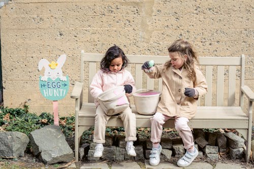 Free Full body positive cute diverse girls in warm clothes playing with Easter eggs and sitting on bench in backyard Stock Photo