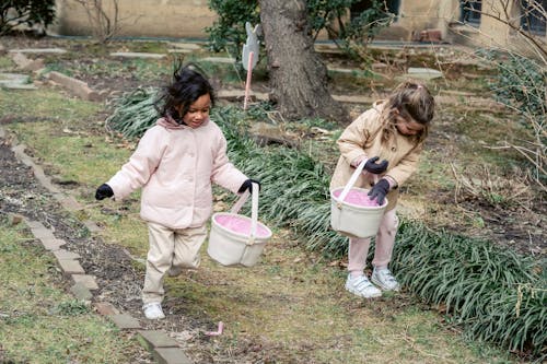 Free Full body glad multiracial girls wearing warm clothes carrying fabric baskets and collecting hidden Easter eggs together in spring park Stock Photo