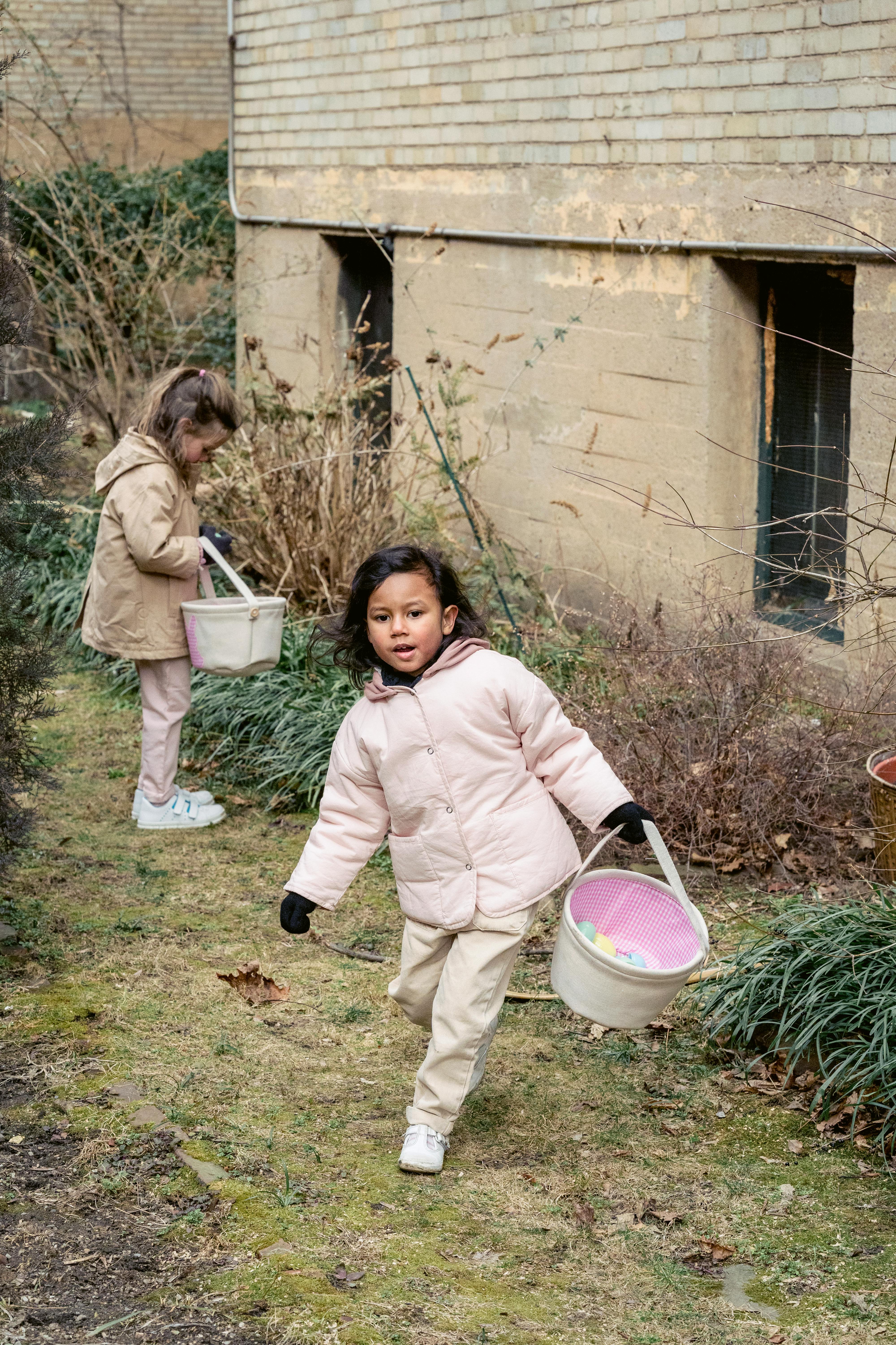 Full length content multiethnic girls in outerwear collecting Easter eggs in fabric baskets in backyard on spring day