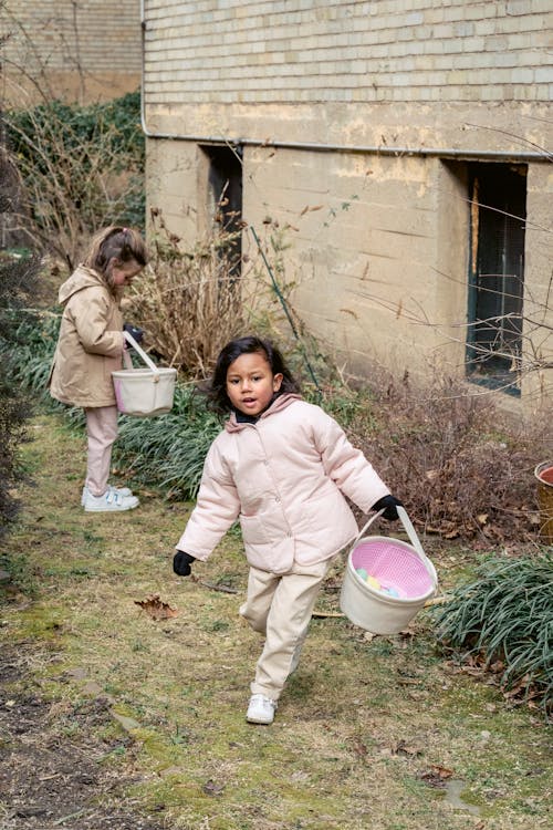 Free Full length content multiethnic girls in outerwear collecting Easter eggs in fabric baskets in backyard on spring day Stock Photo