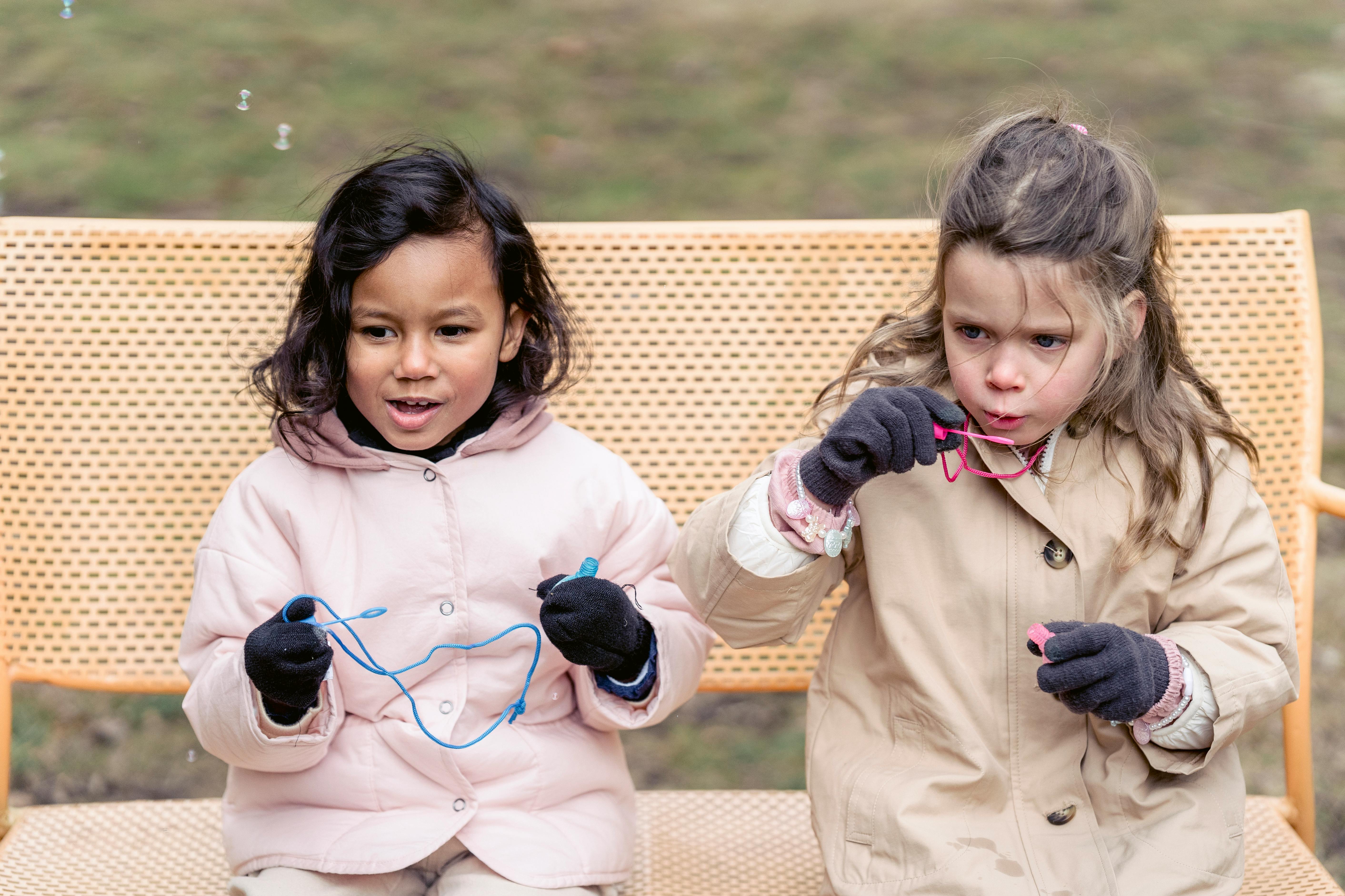 cheerful diverse girls blowing bubbles on park bench