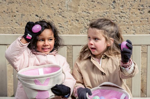 Free Happy diverse girls in warm clothes showing purple Easter eggs and sitting together on bench in courtyard Stock Photo