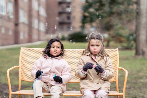 Cute multiracial girls wearing warm outfits playing with soap bubbles on bench in spring park