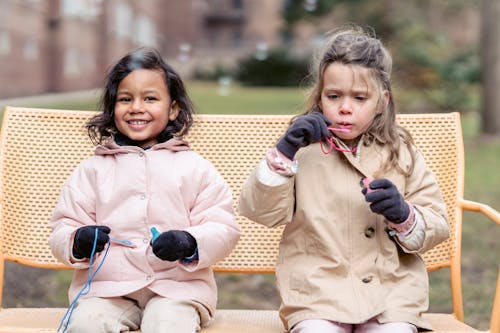 Adorable cheerful diverse girls in outerwear blowing soap bubbles and sitting together on bench in spring garden