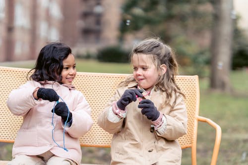 Free Happy multiracial girls wearing warm jackets playing with soap bubbles and looking at each other while sitting on bench in spring garden Stock Photo